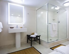 Bright hotel bathroom with illuminated vanity mirror, pedestal sink, glass-enclosed walk-in shower, white towels, bench, neutral tile flooring, and simple contemporary decor.