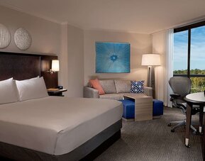 Contemporary king guest room with white bedding, dark headboard, sofa, blue ottomans, work desk and ergonomic chair, floor lamp, colorful wall art, and a large window with a treetop view.