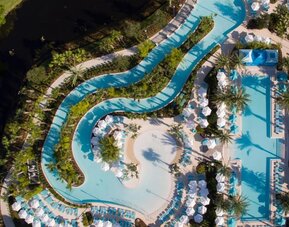 Aerial view of an expansive resort pool complex featuring winding lagoon-style pools, a large main pool, tropical landscaping, palm trees, lounge chairs, umbrellas, and cabanas.