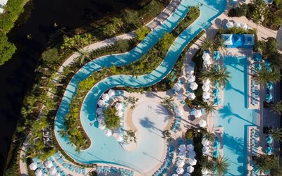 Aerial view of an expansive resort pool complex featuring winding lagoon-style pools, a large main pool, tropical landscaping, palm trees, lounge chairs, umbrellas, and cabanas.