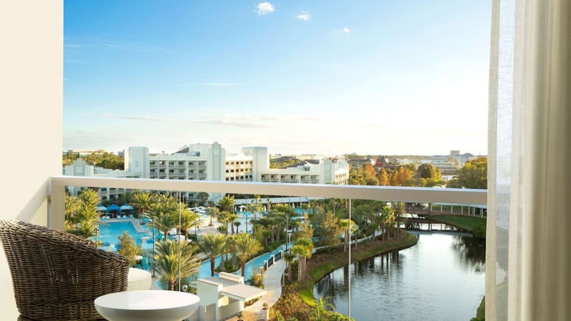 Private hotel balcony with wicker chair and small table overlooking palm-lined resort pools, landscaped grounds, a winding lagoon, and surrounding buildings beneath a bright blue sky.