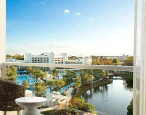 Private hotel balcony with wicker chair and small table overlooking palm-lined resort pools, landscaped grounds, a winding lagoon, and surrounding buildings beneath a bright blue sky.