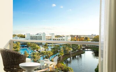 Private hotel balcony with wicker chair and small table overlooking palm-lined resort pools, landscaped grounds, a winding lagoon, and surrounding buildings beneath a bright blue sky.