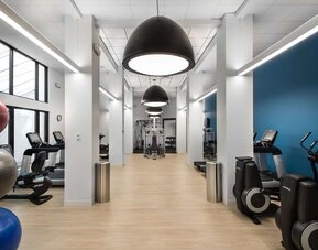 Spacious hotel fitness center with treadmills, elliptical machines, strength-training equipment, exercise balls, and weights beneath oversized pendant lights and a bright contemporary interior.