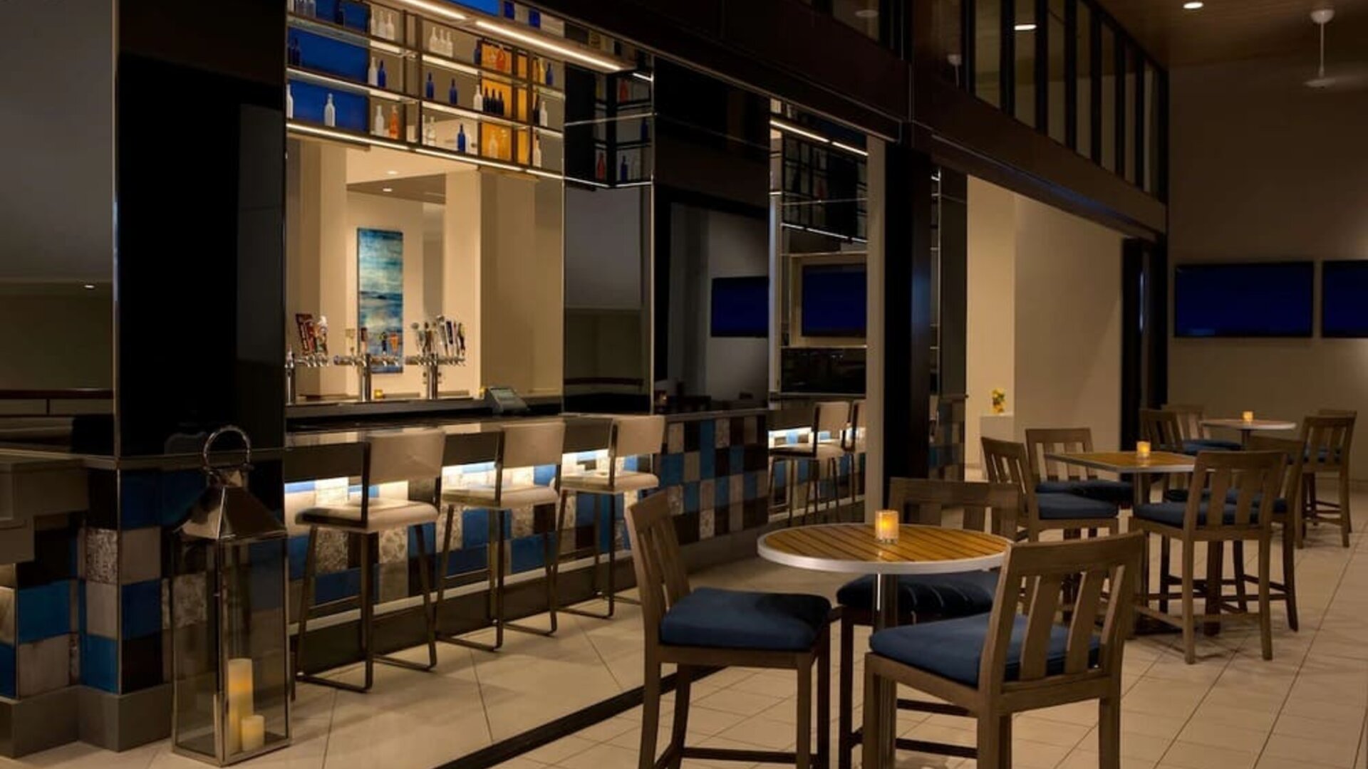 Contemporary hotel bar with illuminated bottle displays, counter seating, high-top tables and chairs, televisions, decorative lanterns, and warm ambient lighting.