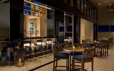 Contemporary hotel bar with illuminated bottle displays, counter seating, high-top tables and chairs, televisions, decorative lanterns, and warm ambient lighting.
