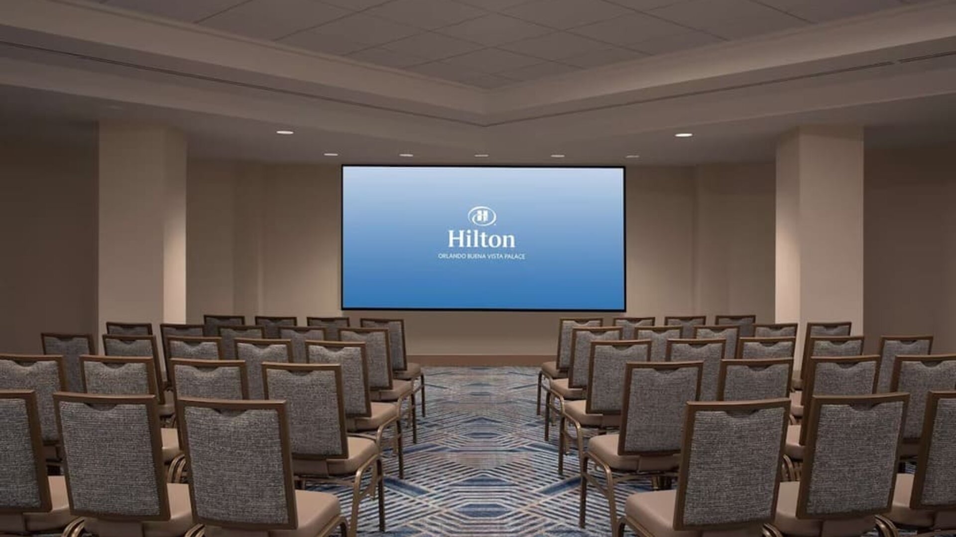 Hotel conference room arranged theater-style with rows of upholstered chairs facing a large presentation screen displaying the Hilton Orlando Buena Vista Palace logo.