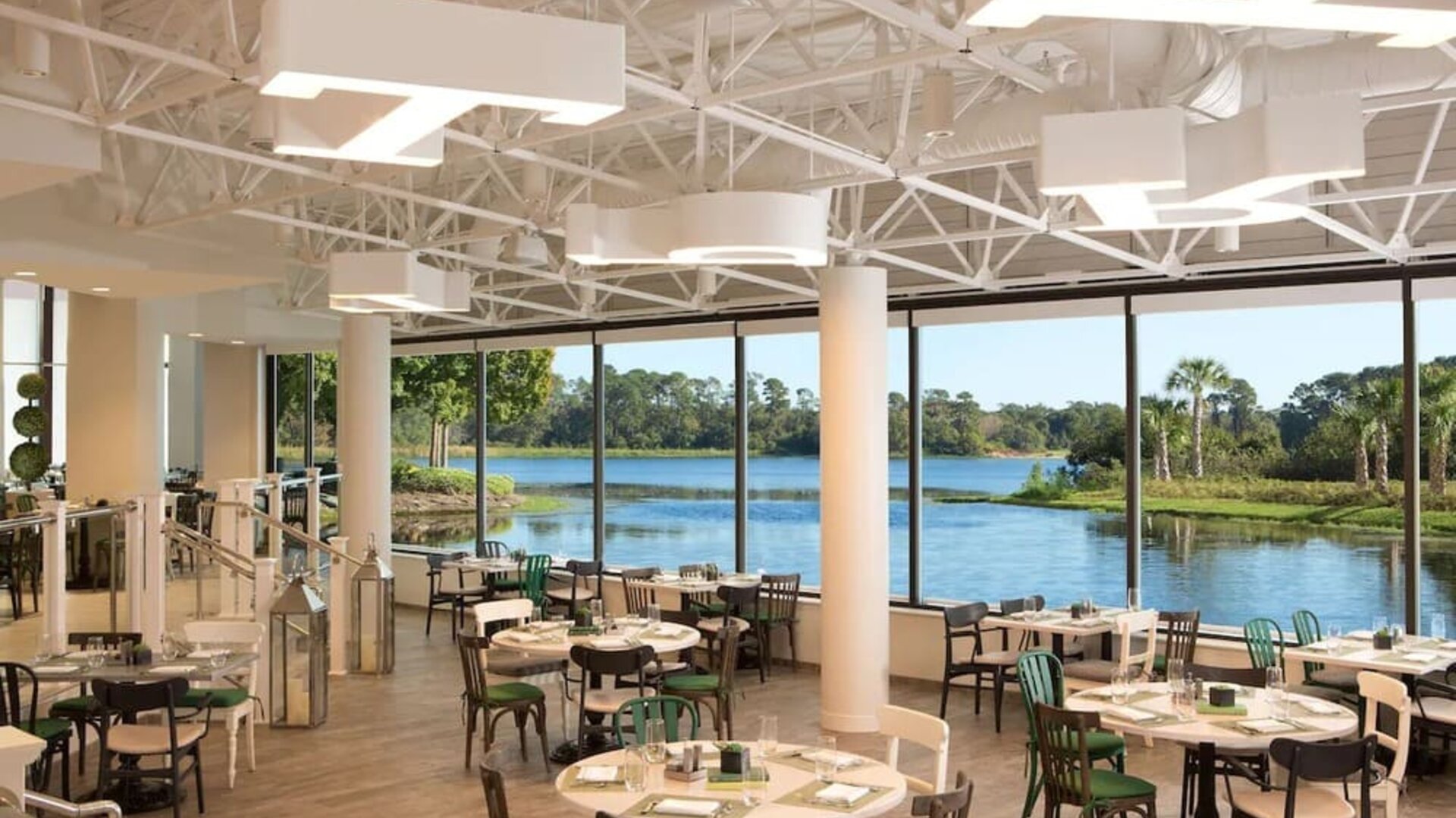 Bright hotel restaurant with round and square dining tables, mixed green, black, and white chairs, modern overhead lighting, and floor-to-ceiling windows overlooking a scenic lagoon and greenery.