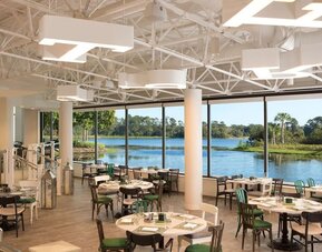 Bright hotel restaurant with round and square dining tables, mixed green, black, and white chairs, modern overhead lighting, and floor-to-ceiling windows overlooking a scenic lagoon and greenery.