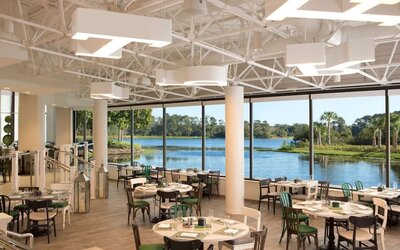 Bright hotel restaurant with round and square dining tables, mixed green, black, and white chairs, modern overhead lighting, and floor-to-ceiling windows overlooking a scenic lagoon and greenery.