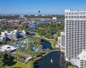 Aerial view of a tall white resort tower beside landscaped grounds, winding waterways, palm-lined swimming pools, and nearby buildings extending across the surrounding area.