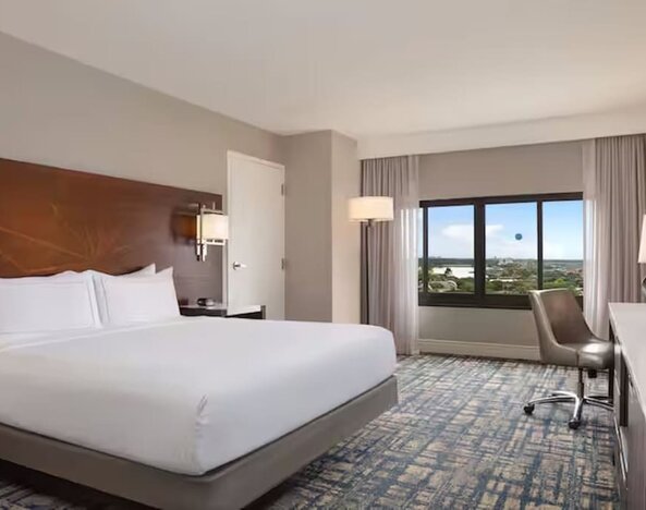 Spacious hotel room with king bed, white linens, large window with an elevated outdoor view, work desk and chair, flat-screen TV, bedside lighting, and patterned carpet.