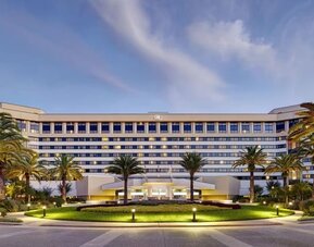 Large multi-story hotel exterior at dusk with illuminated windows and entrance, landscaped circular lawn, mature palm trees, pathways, and exterior lighting.
