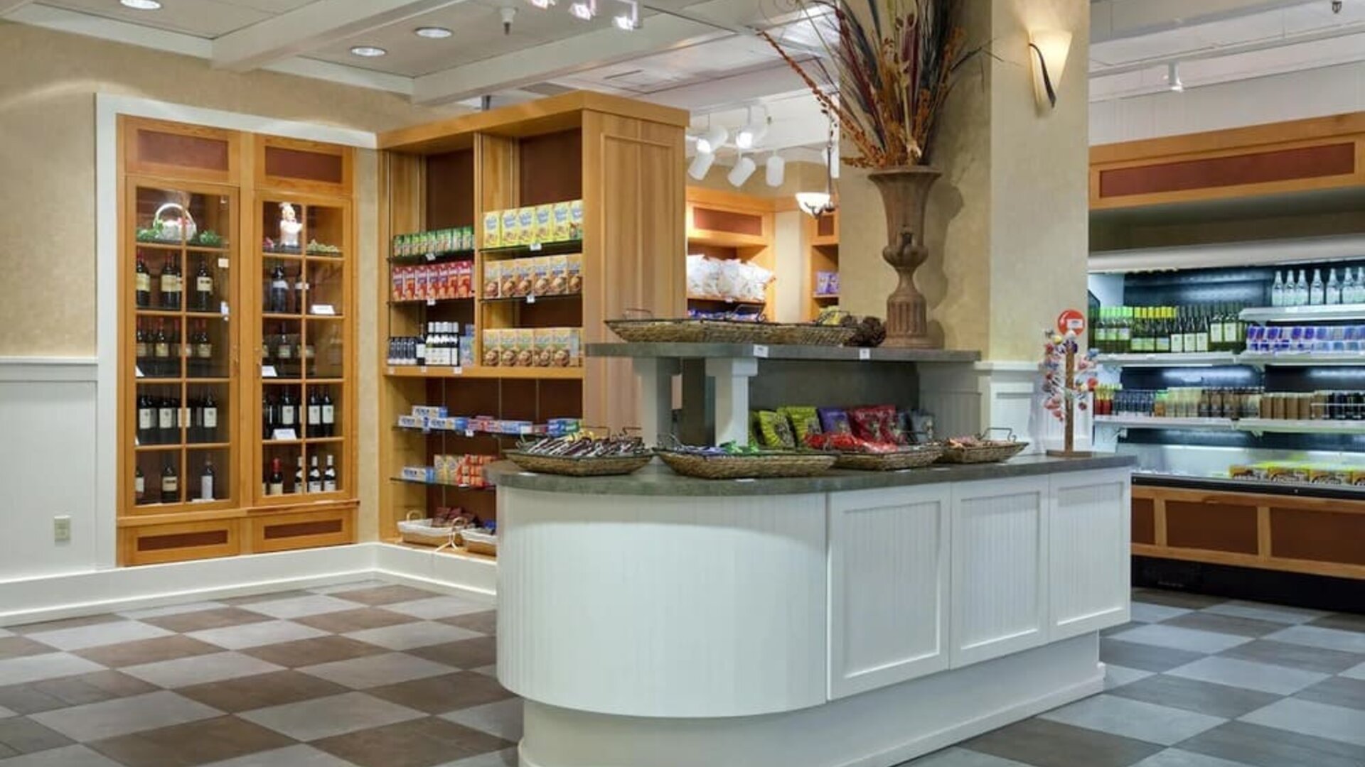 Hotel market with shelves of packaged snacks and beverages, wine display cabinets, refrigerated cases, and a central counter with decorative baskets.