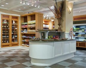 Hotel market with shelves of packaged snacks and beverages, wine display cabinets, refrigerated cases, and a central counter with decorative baskets.