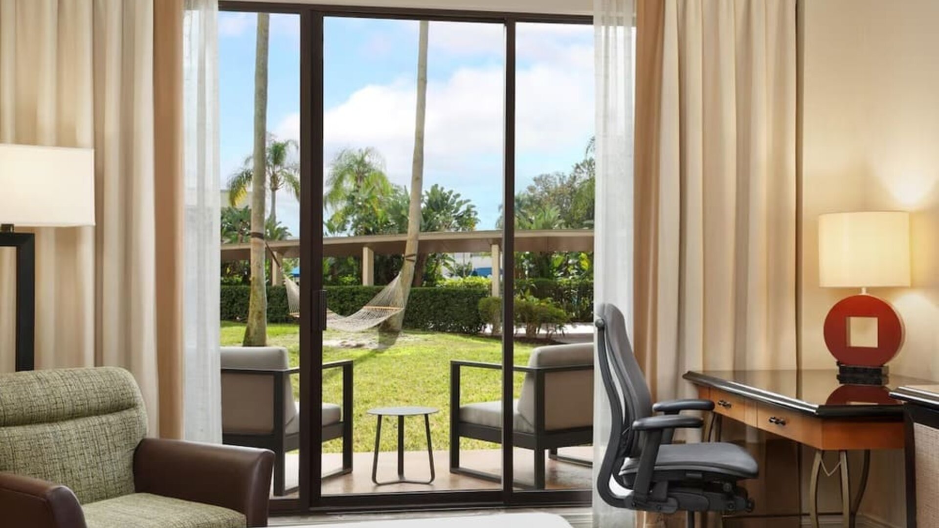 Hotel guest room with desk and ergonomic chair beside sliding glass doors opening to a furnished patio, green lawn, palm trees, and a hammock.