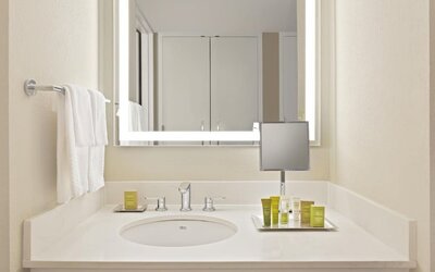 Bright hotel bathroom vanity with white countertop and sink, illuminated rectangular mirror, wall-mounted magnifying mirror, neatly arranged toiletries, and white towels.
