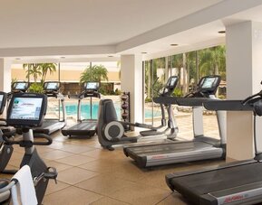 Bright hotel fitness center with treadmills, stationary bikes, elliptical machines, and other cardio equipment overlooking an outdoor swimming pool through large windows.