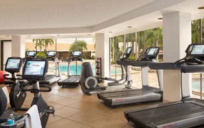 Bright hotel fitness center with treadmills, stationary bikes, elliptical machines, and other cardio equipment overlooking an outdoor swimming pool through large windows.