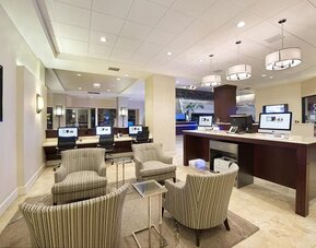 Spacious hotel business center with multiple desktop computers, a printer station, striped lounge chairs, small tables, and modern pendant lighting.