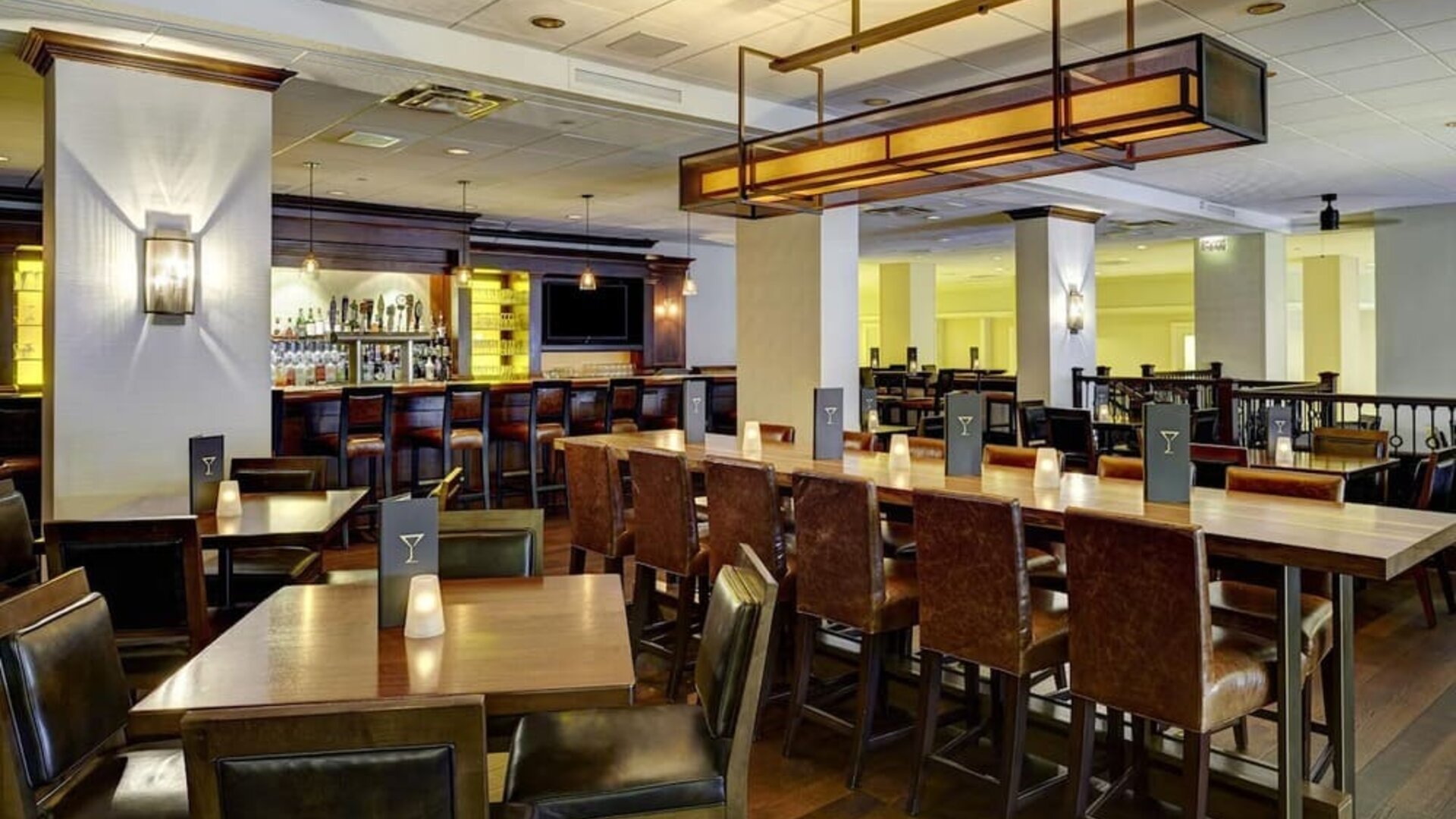 Warm hotel bar and dining area with wood tables, leather chairs, candle-style table lights, high-top communal seating, and a fully stocked bar in the background.