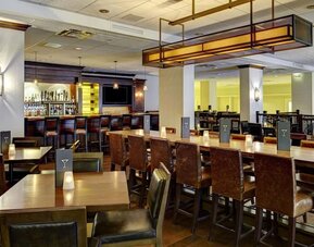 Warm hotel bar and dining area with wood tables, leather chairs, candle-style table lights, high-top communal seating, and a fully stocked bar in the background.