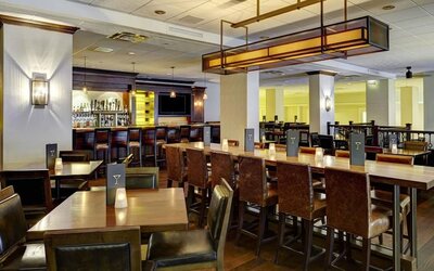 Warm hotel bar and dining area with wood tables, leather chairs, candle-style table lights, high-top communal seating, and a fully stocked bar in the background.