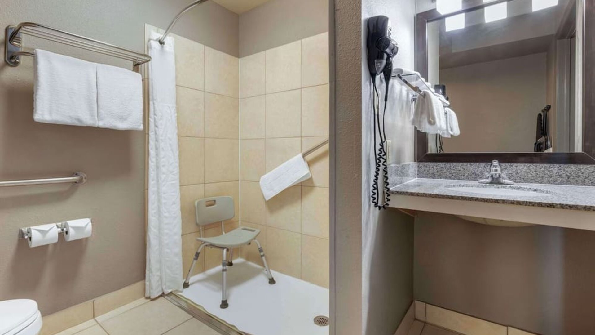 Accessible guest bathroom featuring a roll-in shower with shower chair, grab bars, vanity, mirror, and fresh towels. Best Western McCarran Inn.