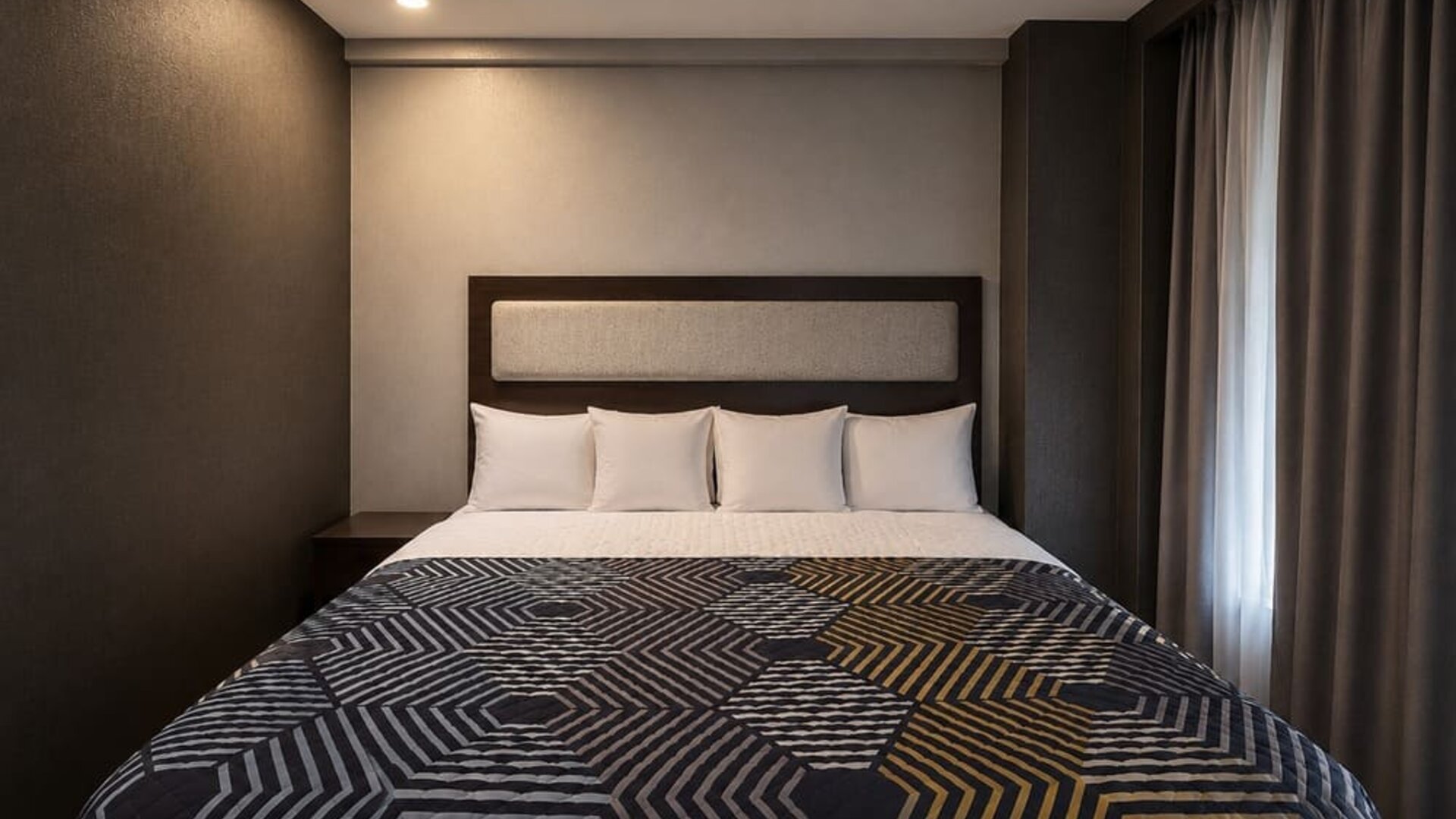 Contemporary king guest room with white pillows, upholstered headboard, dark walls, floor-length curtains, and a bold geometric-patterned bedspread.
