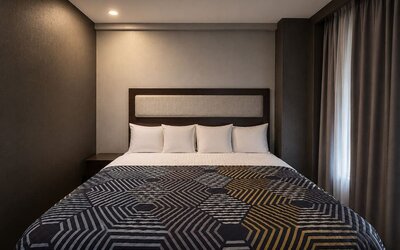 Contemporary king guest room with white pillows, upholstered headboard, dark walls, floor-length curtains, and a bold geometric-patterned bedspread.