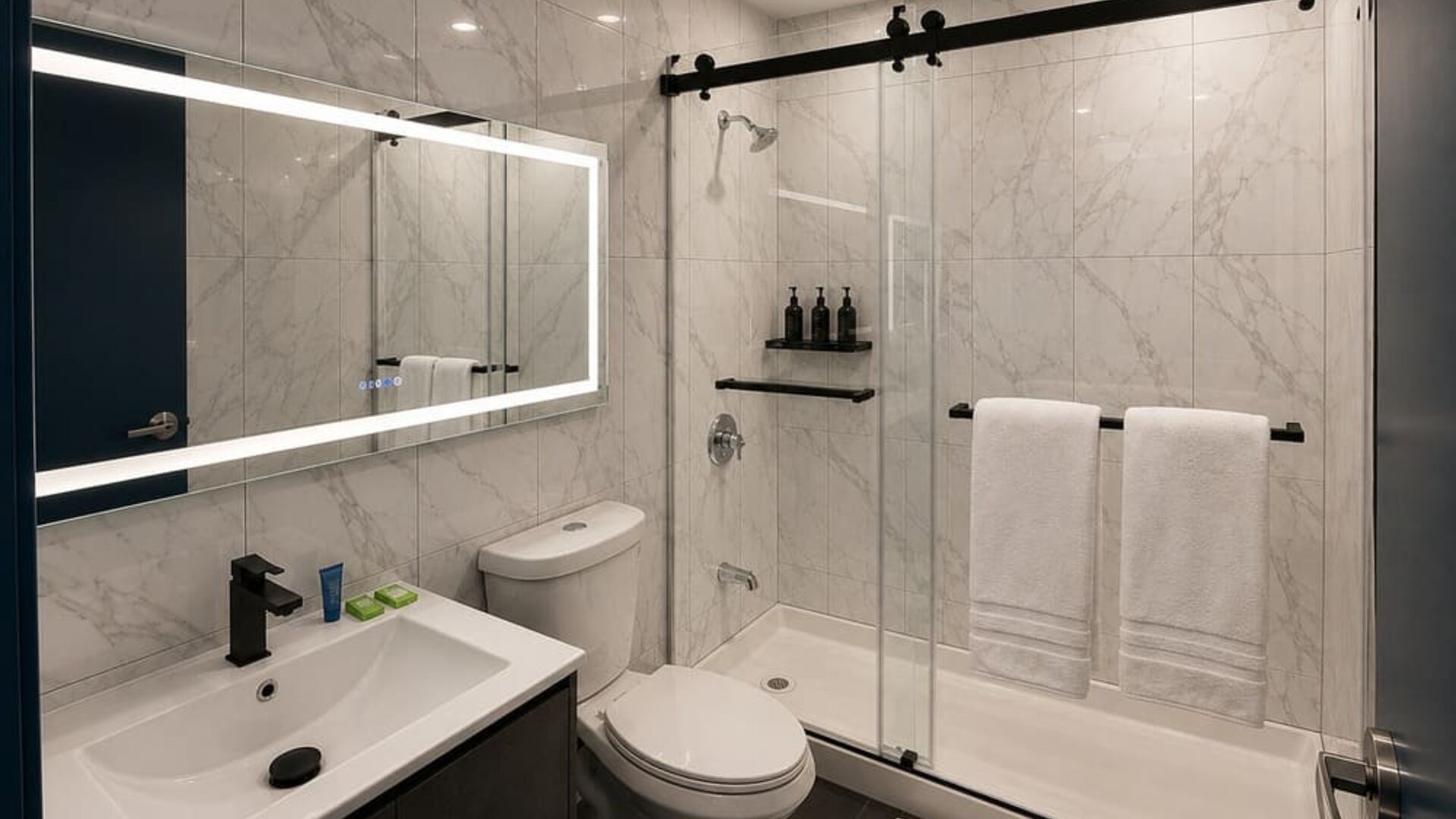 Bright modern bathroom with marble-look tile walls, illuminated mirror, dark vanity, toilet, and glass shower enclosure with black hardware and white towels.