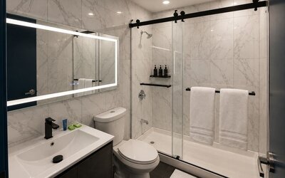 Bright modern bathroom with marble-look tile walls, illuminated mirror, dark vanity, toilet, and glass shower enclosure with black hardware and white towels.
