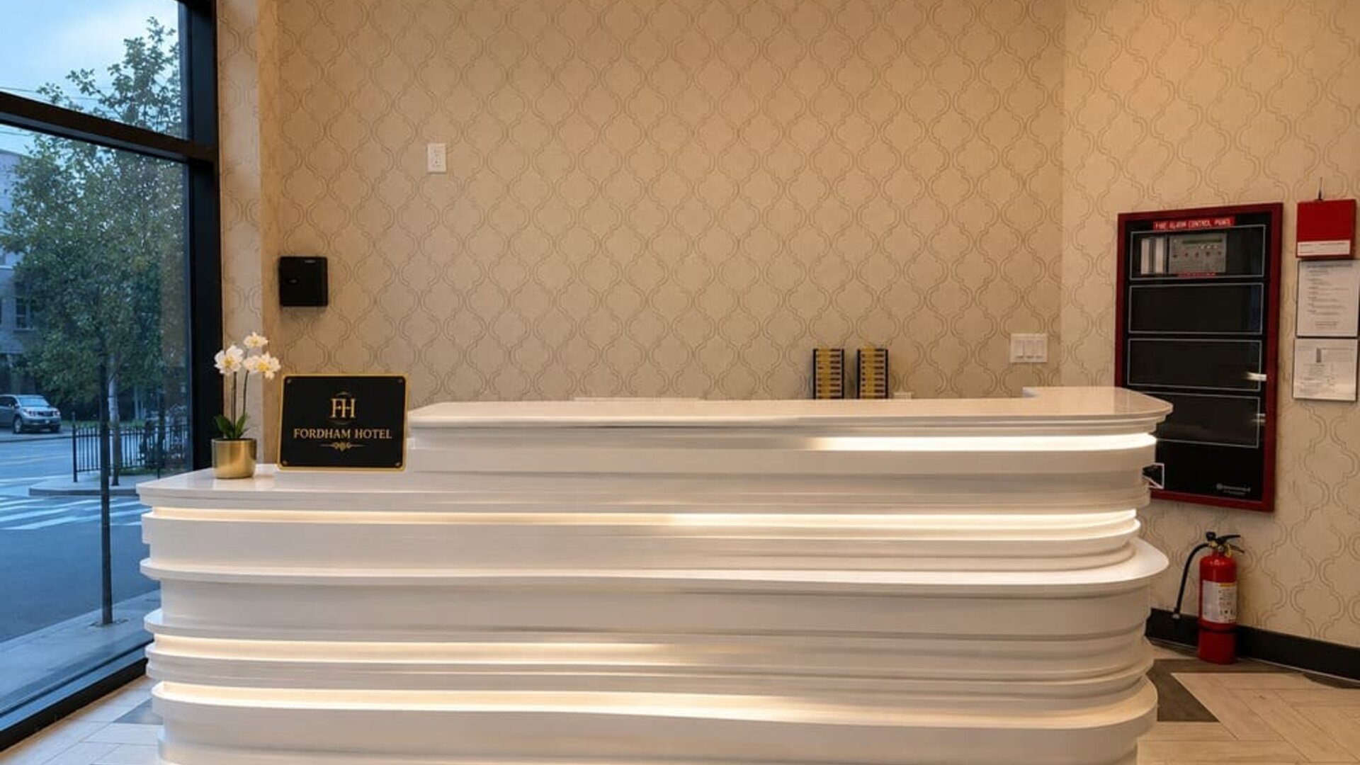 Fordham Hotel reception area with a sculpted white illuminated front desk, patterned beige walls, large street-facing windows, and hotel signage.