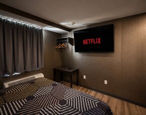 Dark contemporary guest room with geometric-patterned king bed, wall-mounted television displaying Netflix, blackout curtains, open hanging rack, and small table.