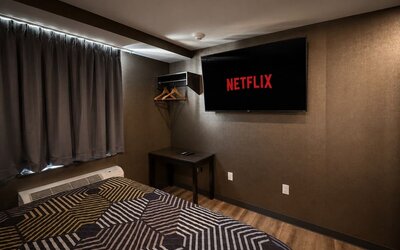 Dark contemporary guest room with geometric-patterned king bed, wall-mounted television displaying Netflix, blackout curtains, open hanging rack, and small table.