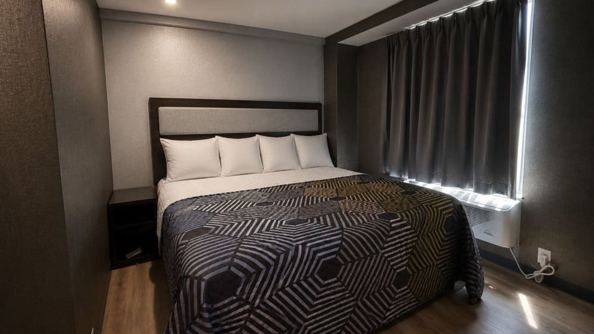 Compact modern king room with white pillows, geometric black-and-gold bedspread, dark walls and curtains, wood-look flooring, and a window air-conditioning unit.