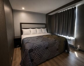 Compact modern king room with white pillows, geometric black-and-gold bedspread, dark walls and curtains, wood-look flooring, and a window air-conditioning unit.