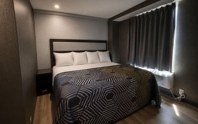 Compact modern king room with white pillows, geometric black-and-gold bedspread, dark walls and curtains, wood-look flooring, and a window air-conditioning unit.