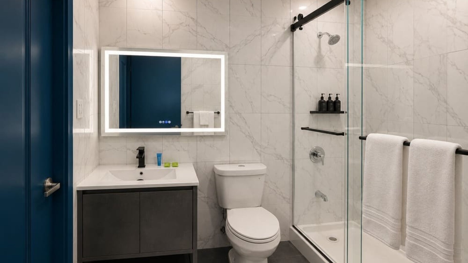 Modern hotel bathroom with marble-look wall tiles, illuminated mirror, white vanity, toilet, and glass-enclosed shower with black fixtures and white towels.