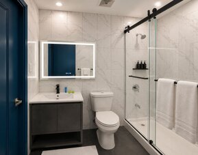 Modern hotel bathroom with marble-look wall tiles, illuminated mirror, white vanity, toilet, and glass-enclosed shower with black fixtures and white towels.