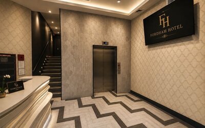 Fordham Hotel lobby with curved white reception desk, elevator, illuminated staircase, patterned tile flooring, decorative walls, and large hotel sign.