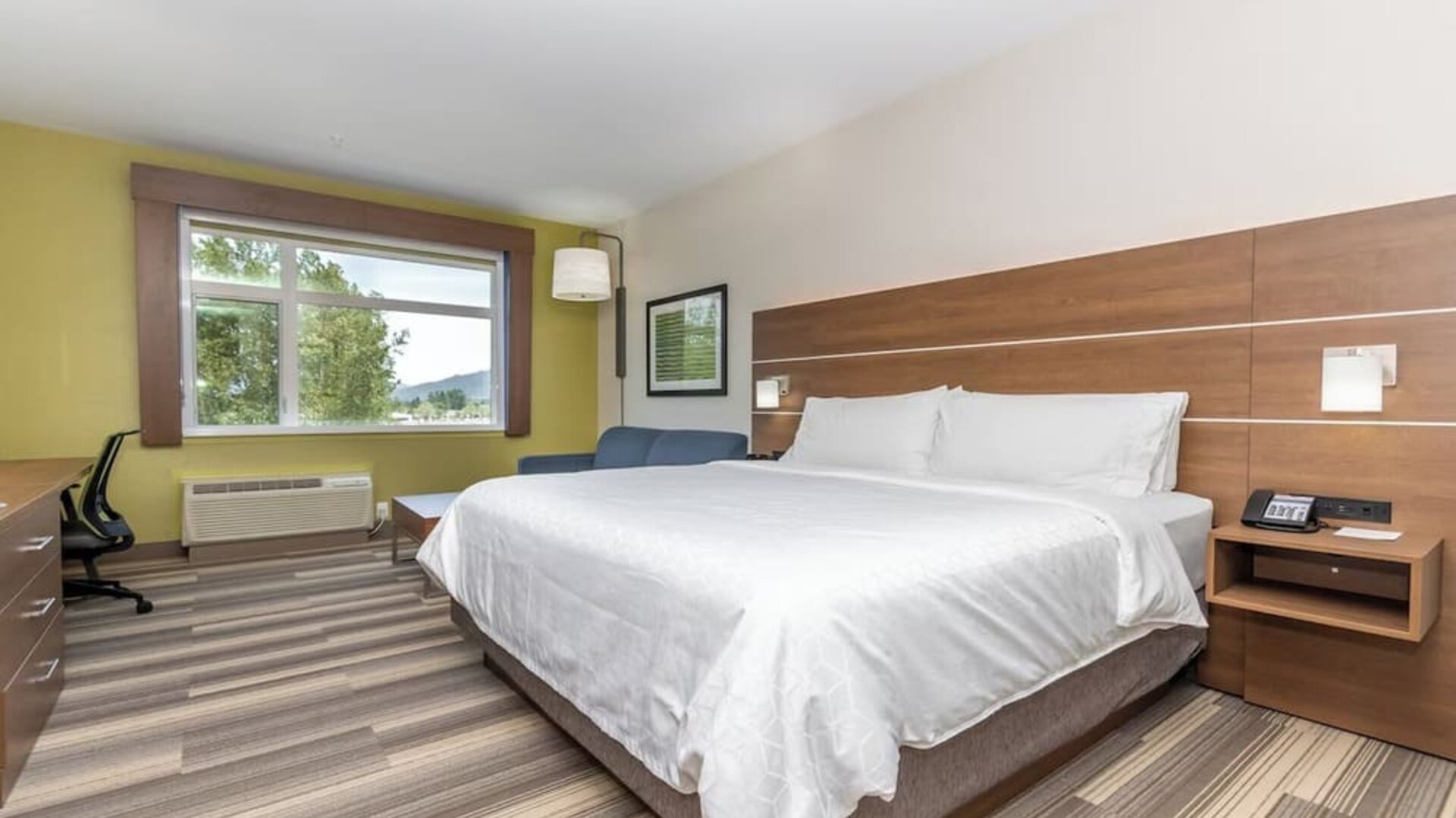Spacious hotel guest room with a king-size bed, white linens, wood headboard, bedside table, work area, seating, and large window with an outdoor view.