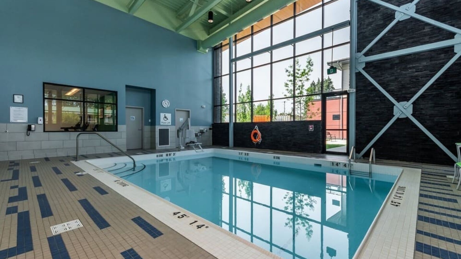 Indoor hotel swimming pool with a tiled deck, floor-to-ceiling windows, accessibility lift, safety equipment, and contemporary high-ceiling design.