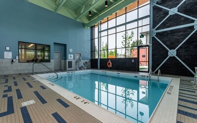 Indoor hotel swimming pool with a tiled deck, floor-to-ceiling windows, accessibility lift, safety equipment, and contemporary high-ceiling design.