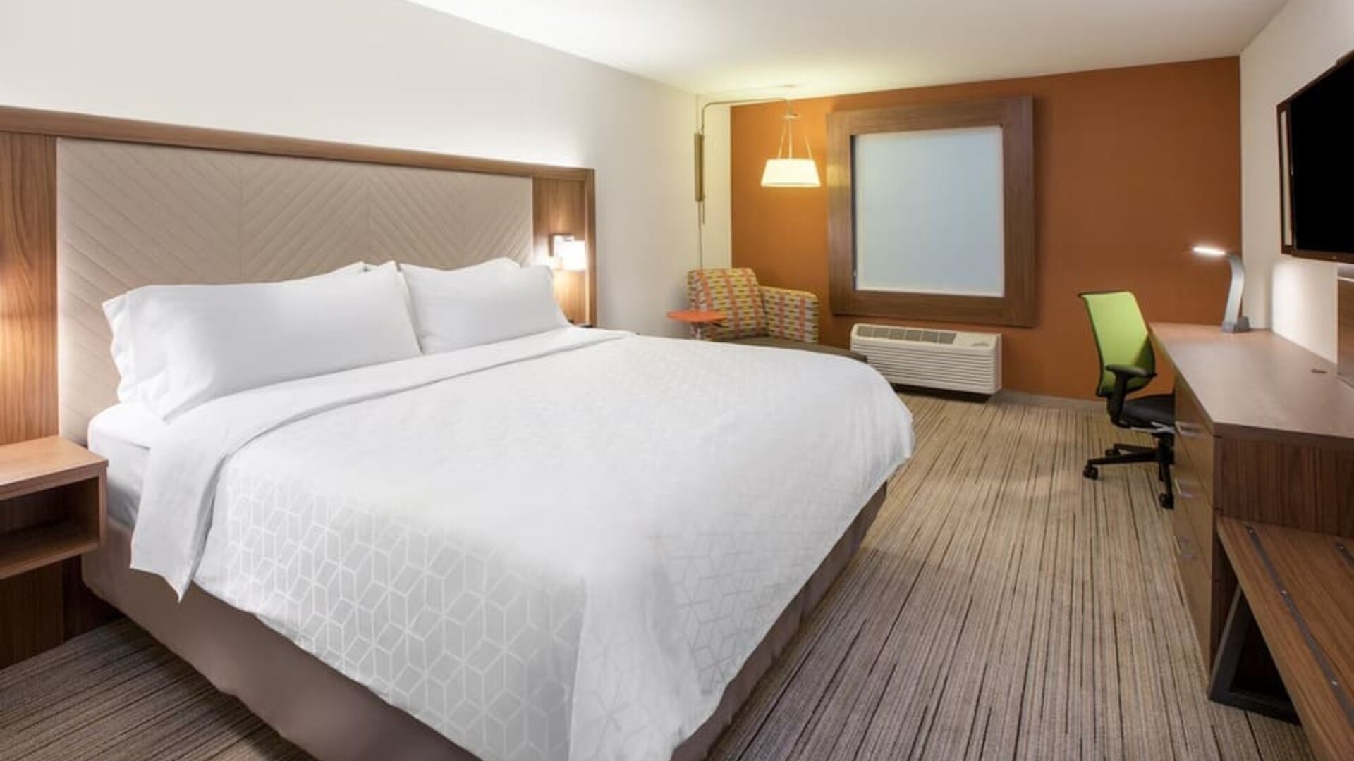 Modern hotel room with a king-size bed, white bedding, work desk and chair, wall-mounted TV, accent chair, and warm wood furnishings.