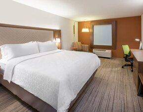Modern hotel room with a king-size bed, white bedding, work desk and chair, wall-mounted TV, accent chair, and warm wood furnishings.
