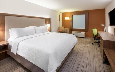 Modern hotel room with a king-size bed, white bedding, work desk and chair, wall-mounted TV, accent chair, and warm wood furnishings.