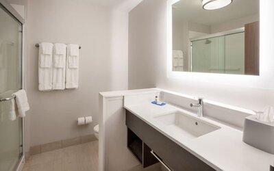 Bright hotel bathroom with a large illuminated mirror, white vanity and sink, glass-enclosed shower, toilet, and neatly arranged white towels.