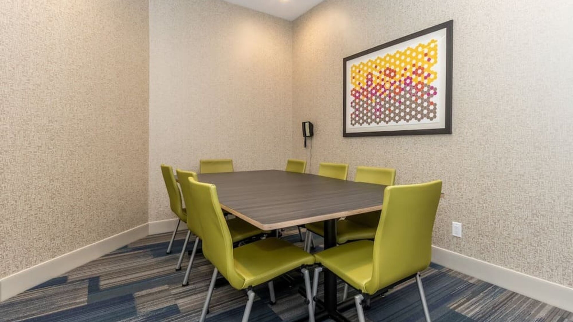 Private hotel meeting room with a rectangular conference table, eight green chairs, patterned carpet, wall-mounted telephone, and colorful framed artwork.