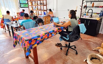 Spacious coworking area with several people working on laptops at colorful communal tables, assorted chairs, wooden display shelving, kitchenette, and a dog resting on the floor.